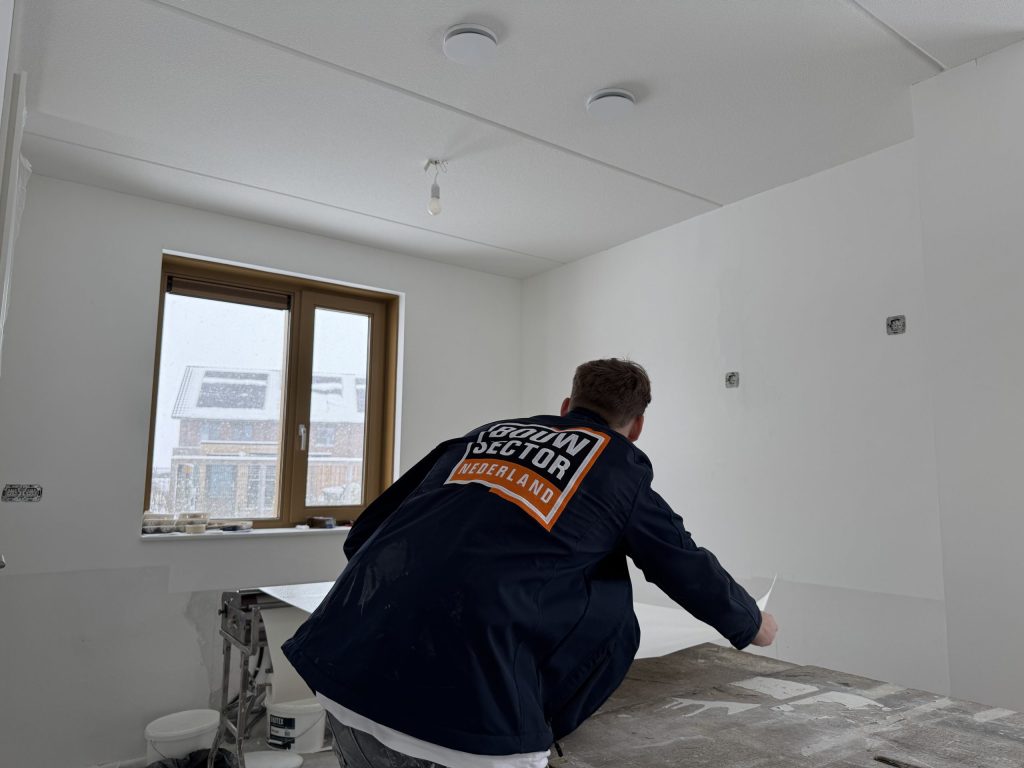 Behanger in donkere werkjas legt een baan behang recht op de plaktafel