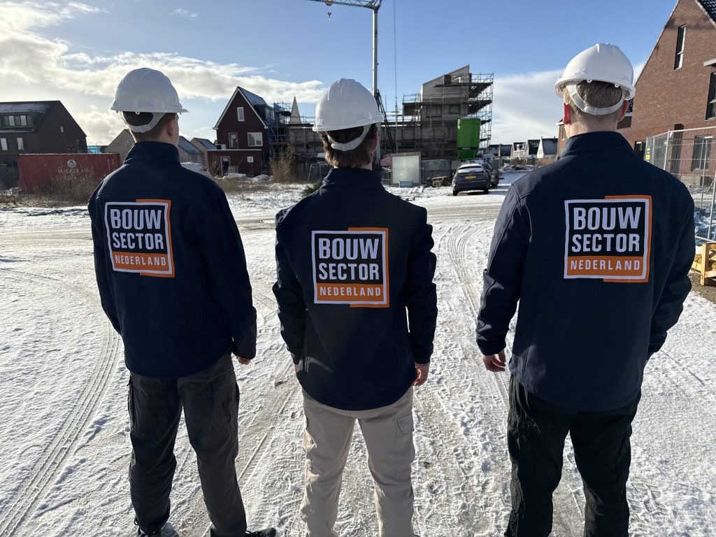 Drie vakmensen van Bouwsector Nederland met witte helm op een besneeuwde bouwplaats met nieuwe woningen